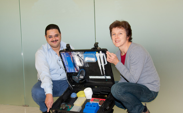 The photo shows two scientists and a suitcase.
